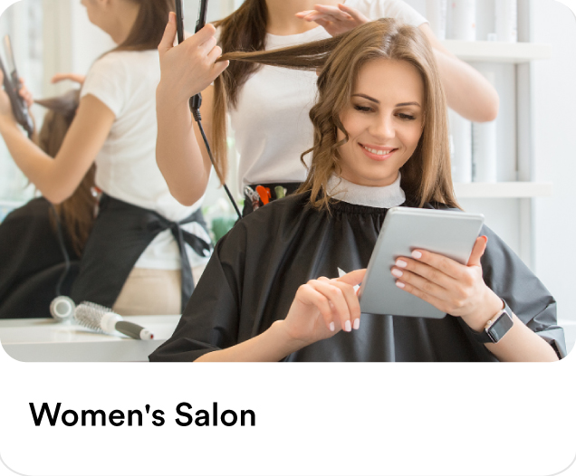 Professional hairstylist styling a woman's hair while she looks at a tablet to illustrate premium beauty services and women's salon at home in the UAE.